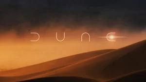 A imagem apresentada é um cenário de deserto, com dunas suavemente iluminadas pelo crepúsculo ou amanhecer, criando um gradiente de cores que vai do laranja ao marrom escuro. No centro da imagem, está a palavra "DUNE" escrita em letras maiúsculas, com um design estilizado e minimalista. O fundo é dominado por um céu escuro, possivelmente com nuvens ou uma tempestade de areia ao longe, contribuindo para a atmosfera misteriosa e árida do ambiente desértico.