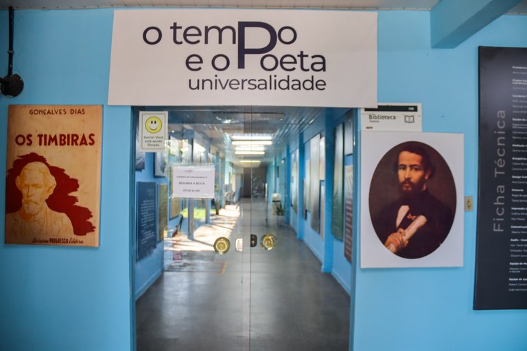 Imagem apresenta a entrada da Biblioteca Central da UFMA com o cartaz exibindo o título da exposição "O tempo e o Poeta". 