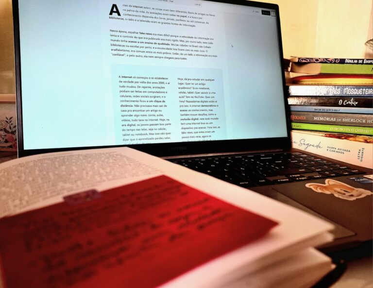 Notebook cinza com livro aberto e marcador vermelho e pilhas de livros representando o aprendizado na era digital.